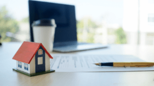a work space with a laptop, coffee cup, pencil and paper, and miniature house